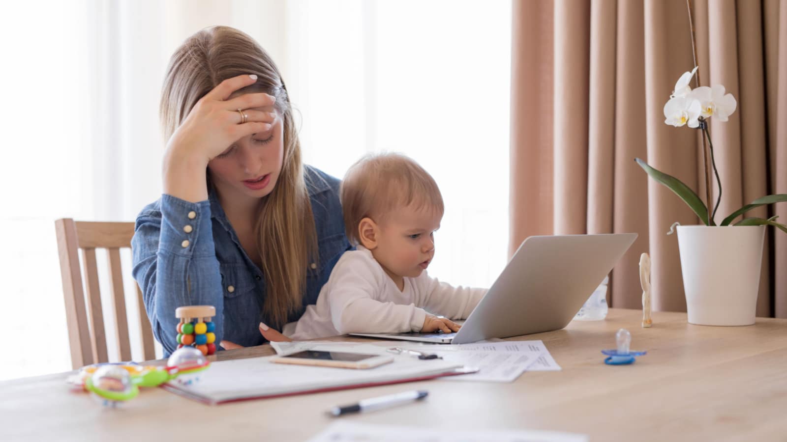 How much does it cost to raise a child? A mom sits stressed at her laptop holding a baby.