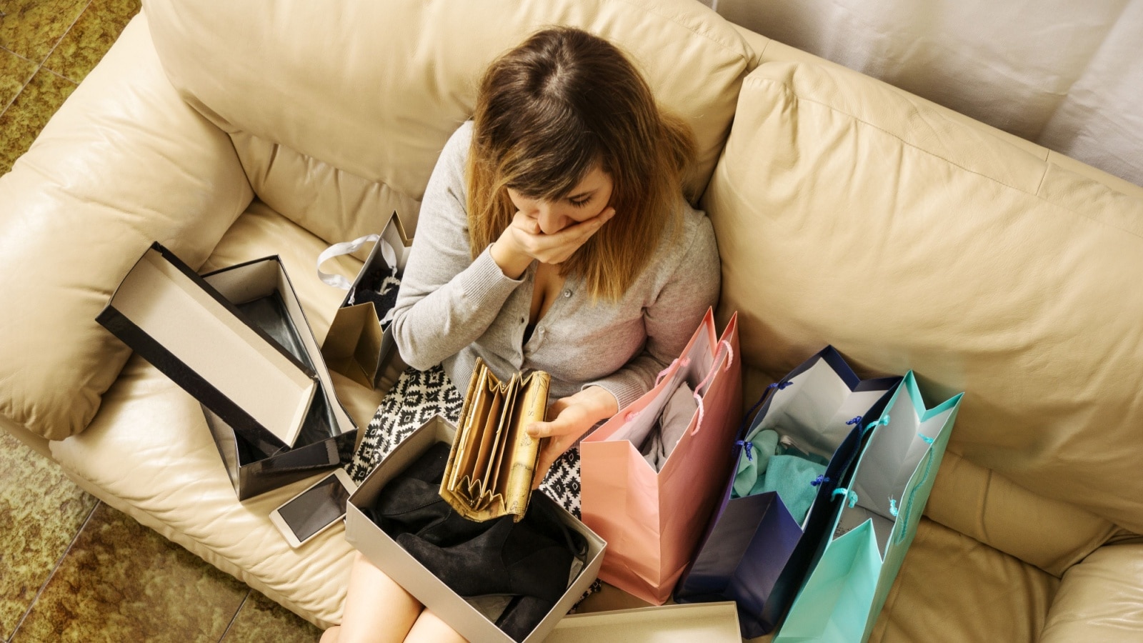 A woman sits on her couch surrounded by shopping bags but gasps as she looks into her empty wallet.