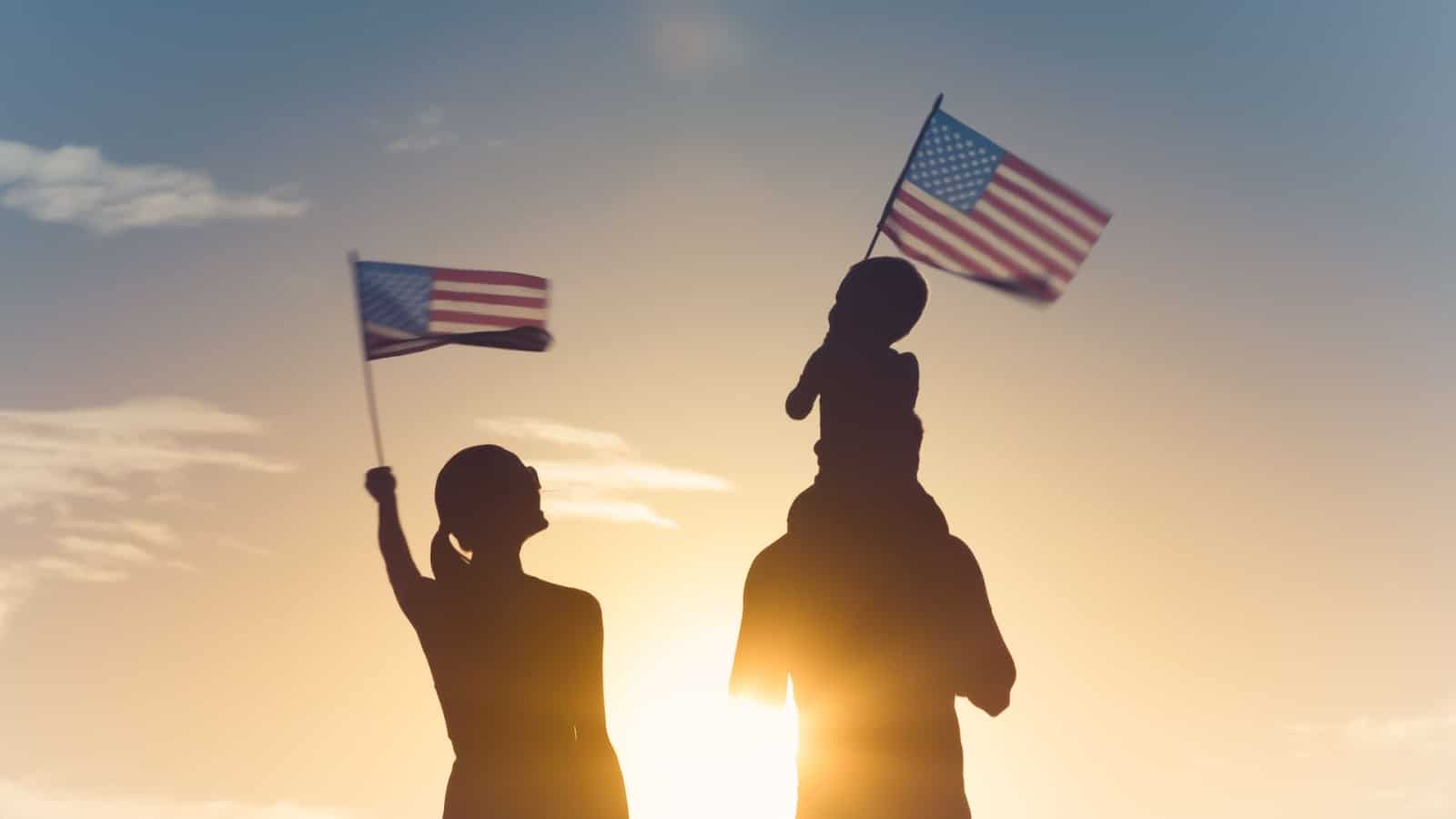 Siloutte of family waving American flags in the ealry morning sun to symbolize the American Dream. The child is sitting on the father's shoulders