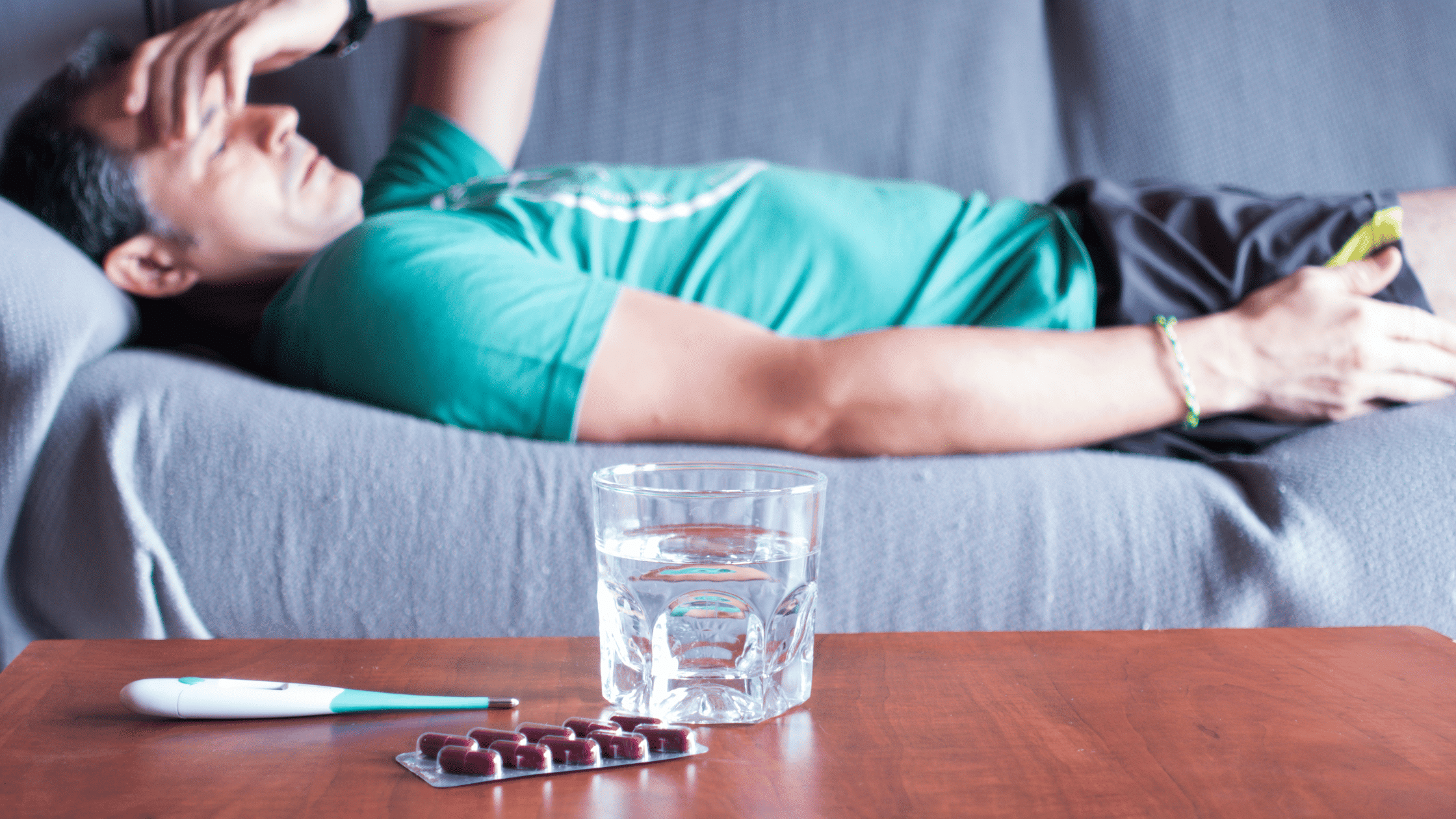man laying on a couch, hand on his head with thermometer and pills on table showcasing he's sick to represent paid sick leave policies
