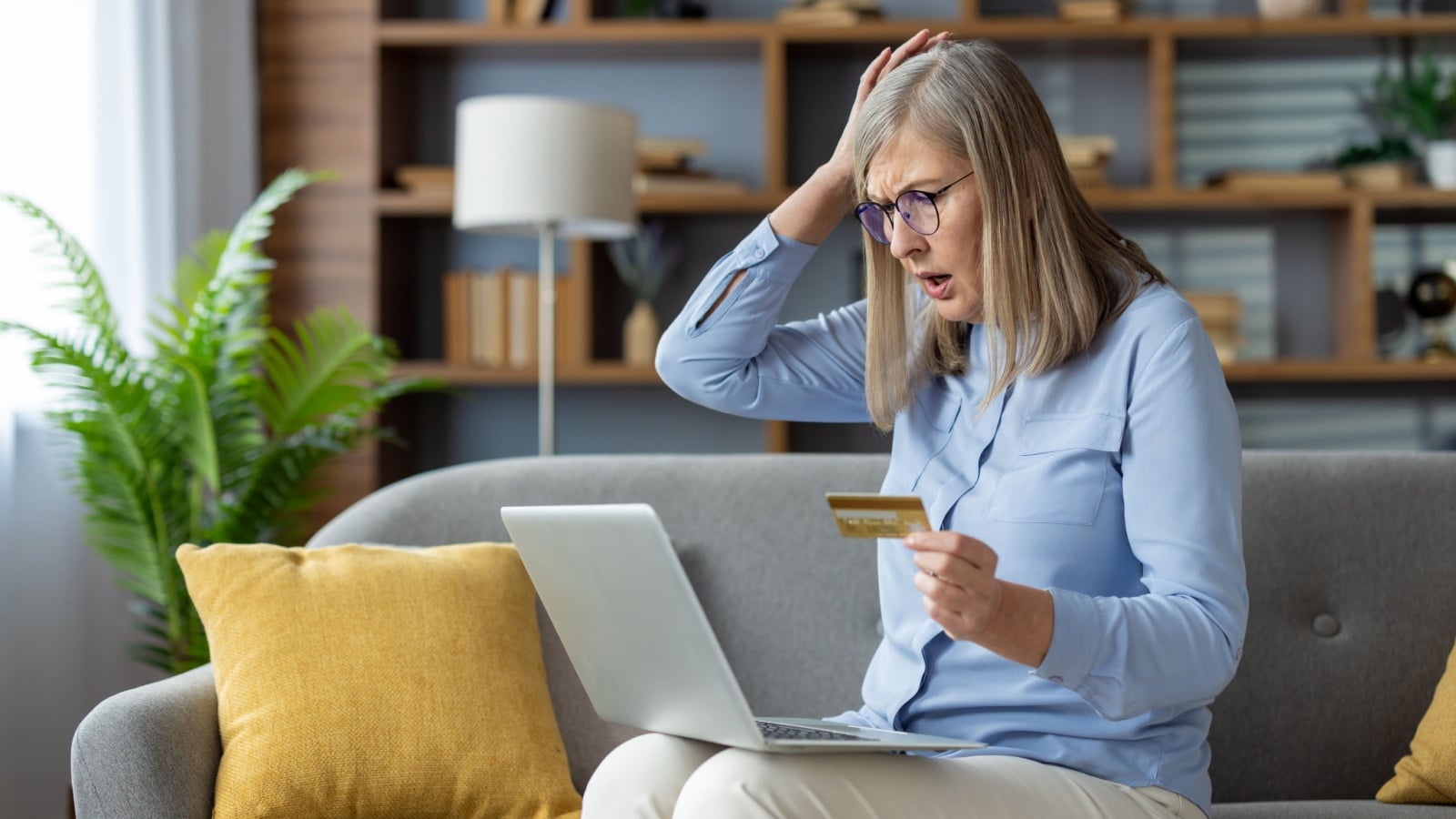What to do if your identity is stolen. A woman sits on the couch, holding a credit card and gasping at the computer. She's a victim of identity theft.