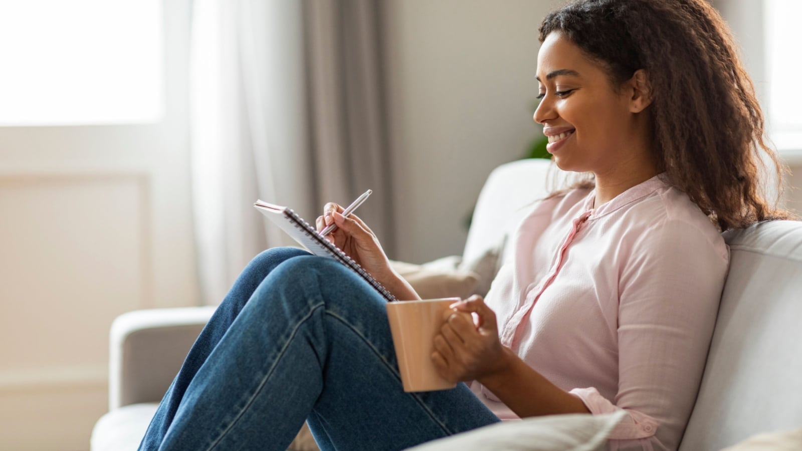 A happy woman sitting on the coach writing in her journal to represent journal prompts.