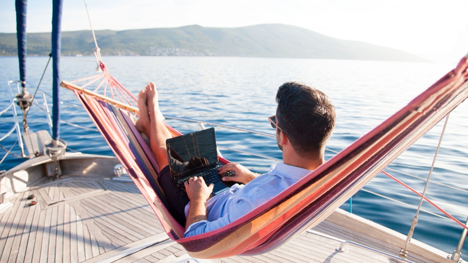 A man works as a digital nomad. He's laying in a hammock on a boat, working on his laptop. 