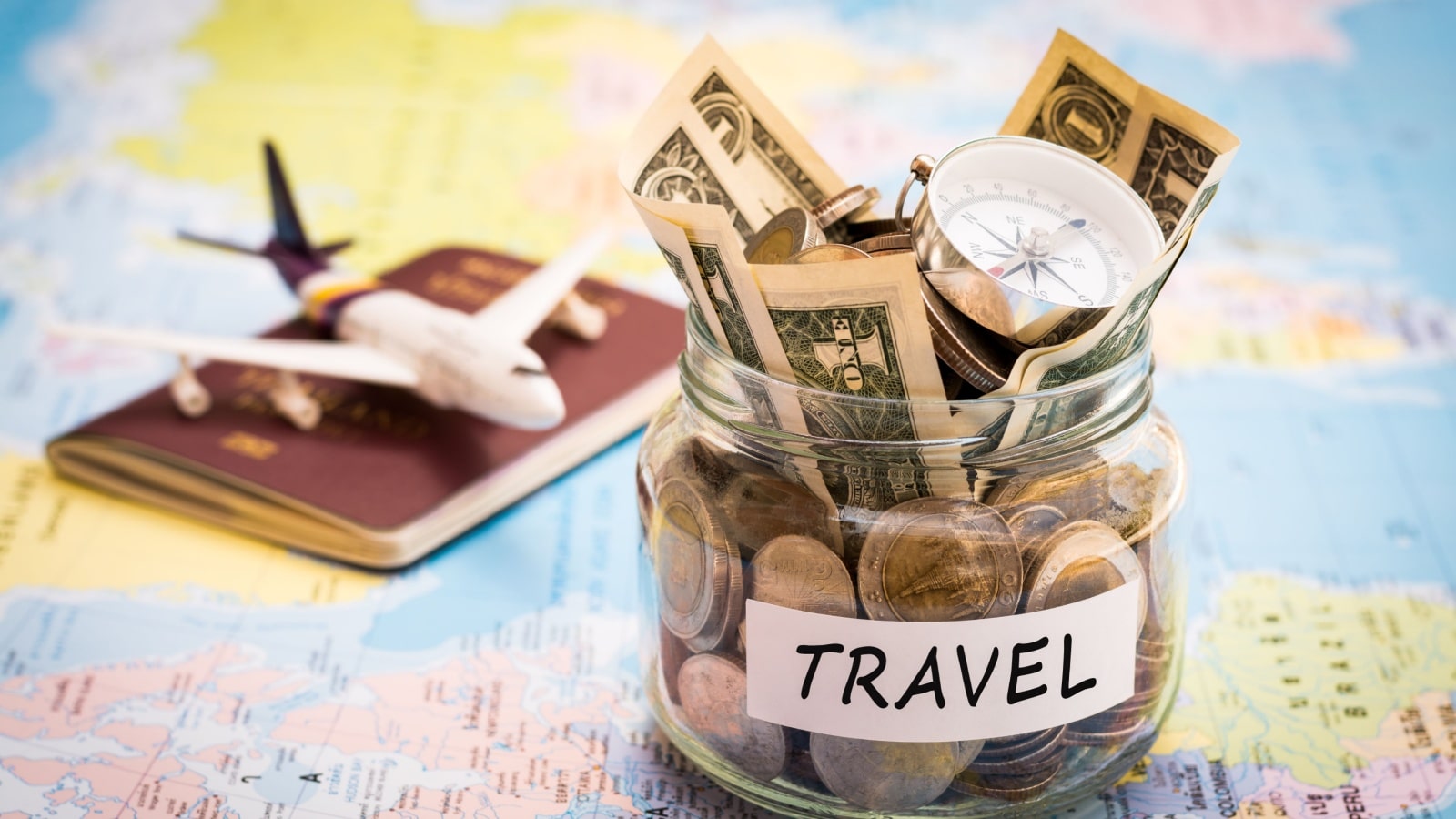 Get paid to travel concept art. Money is in a jar labelled "travel" sitting on a map. There's a passport with a model airplane on it in the background.