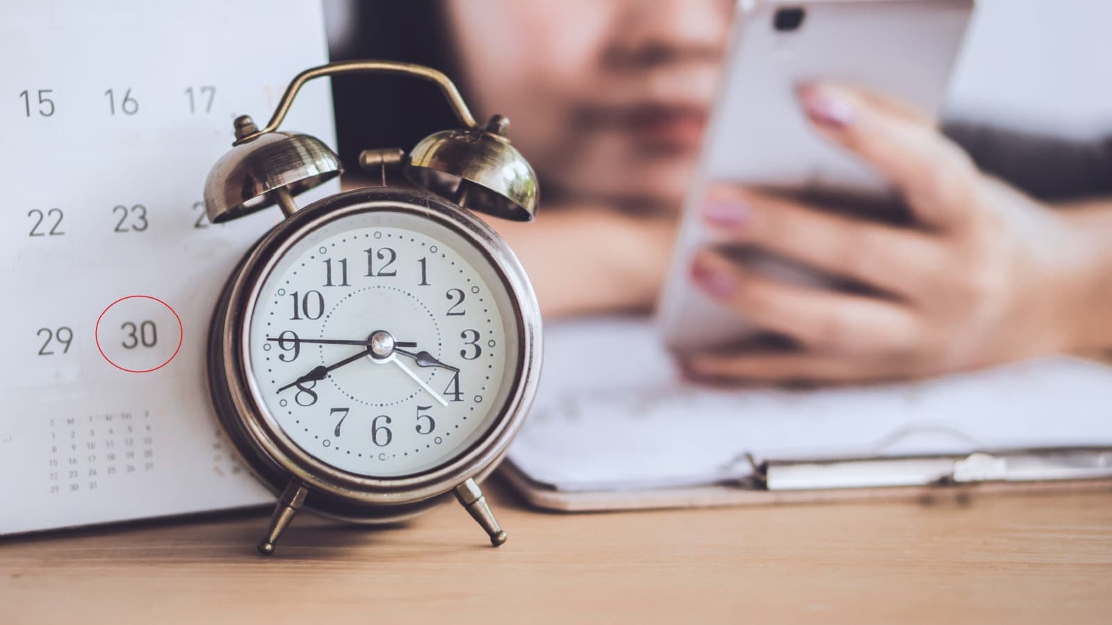 Time wasters aesthetic. An alarm clock sits in front of a calendar, while a woman in the background mindlessly scrolls on her phone wasting time.