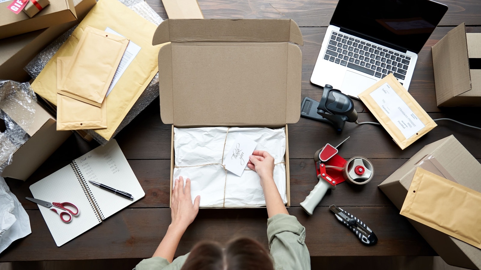 A woman packing up an item for shipping. She's using her desk with a laptop and shipping supplies on it to represent selling stuff online.