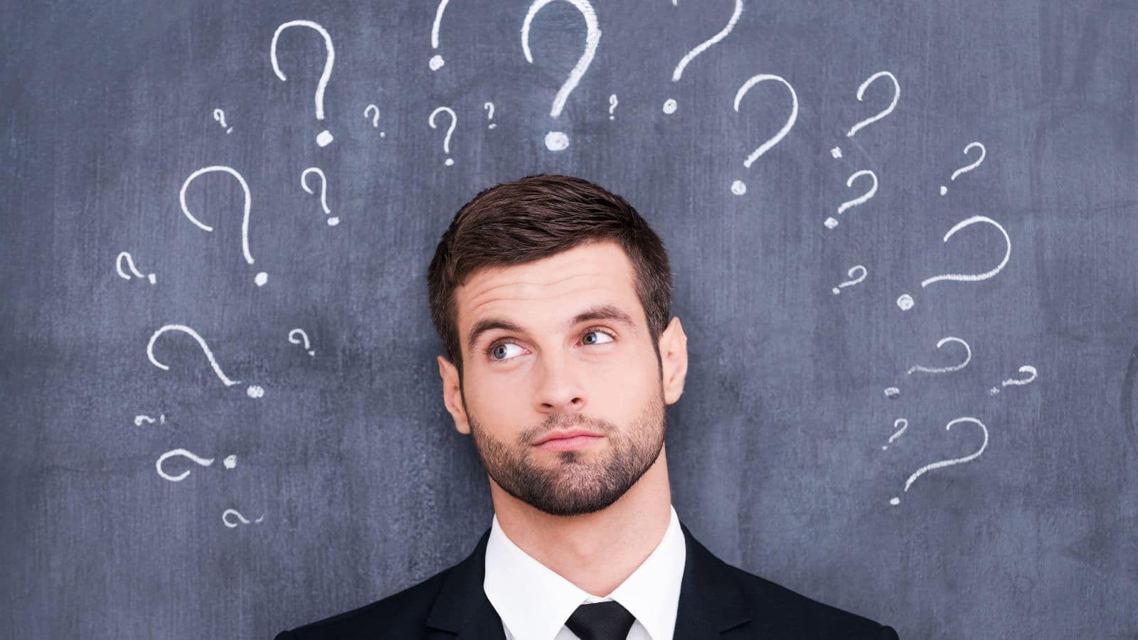 A business man stands in front of a chalkboard, thinking about what to do. The chalkboard has question marks surrounding his head. The image represents the question "what is my purpose in life?"
