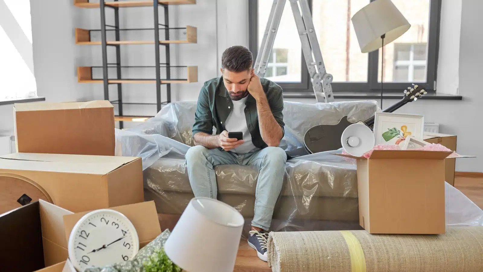 A man sits in the middle of a cluttered mess while getting ready to move to represent a disadvantage of geoarbitrage: that moving is awful.