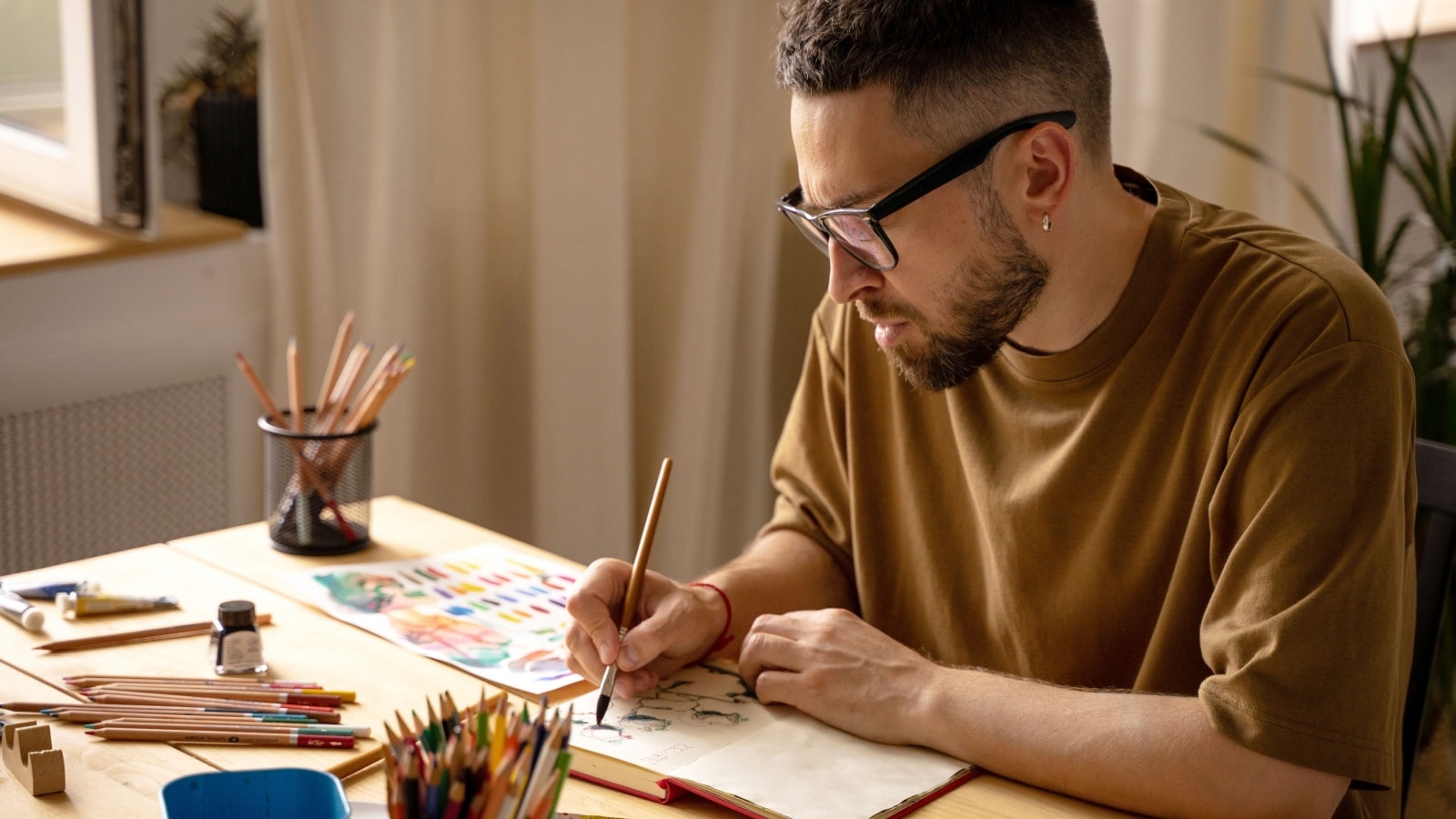 A man sketching in his journal with art journal supplies like pencils and watercolor on his table.