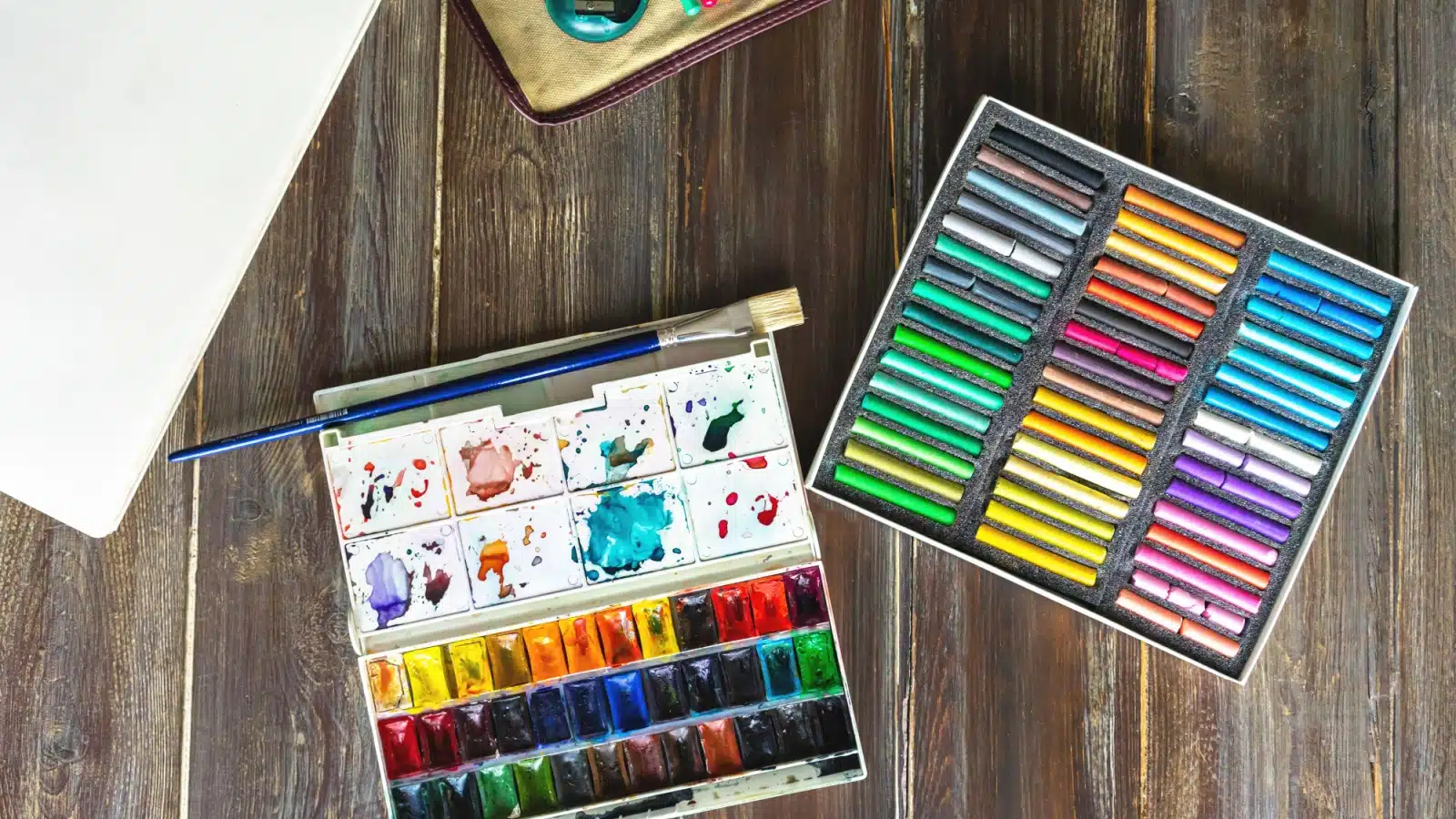 Art supplies for journaling like pastels and watercolor, with part of a notebook. 
