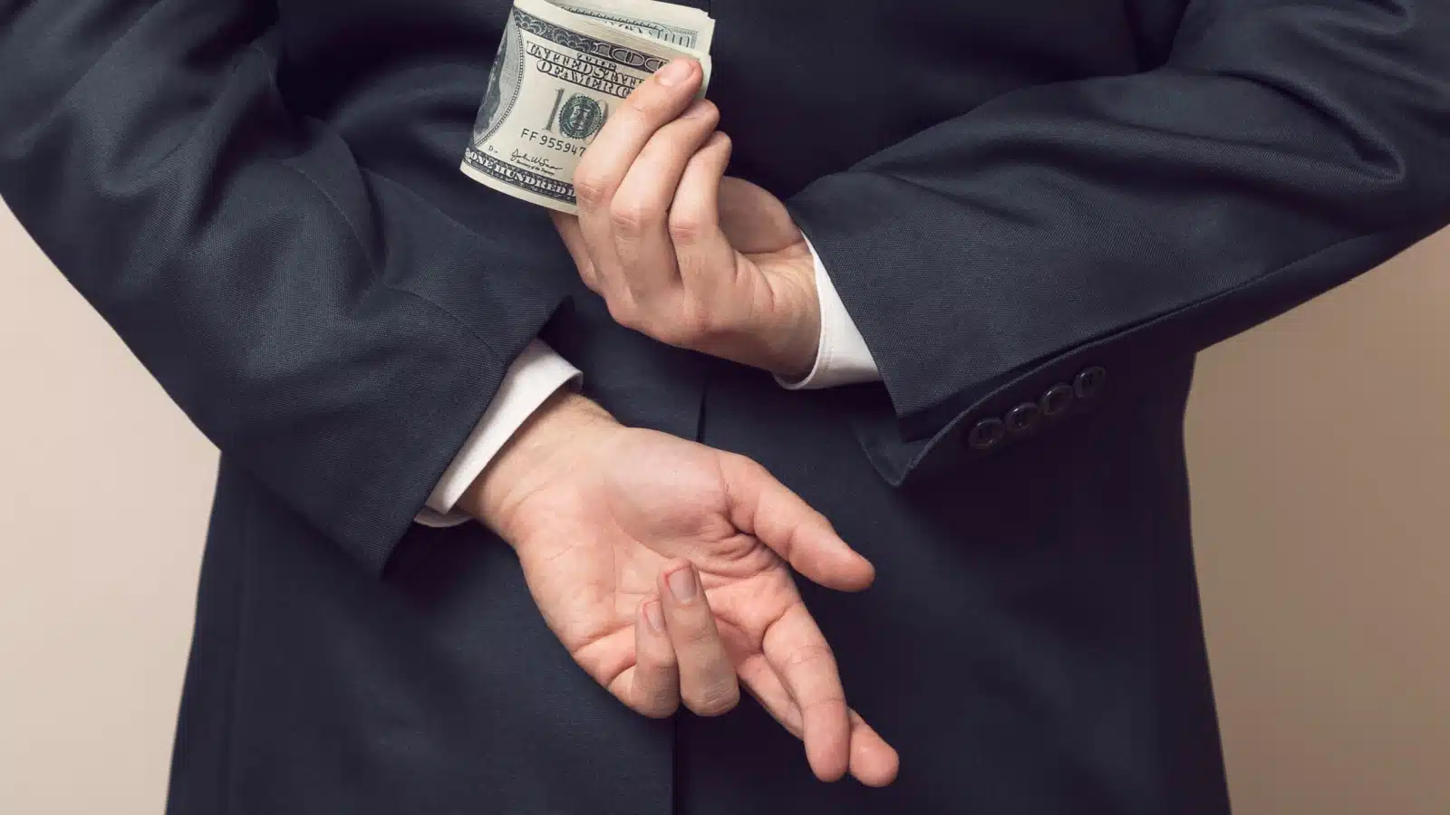 A businessman holds his hands behind his back. He has money in one hand, and his fingers on the other hand are crossed, showing that he's lying and stealing the money. 