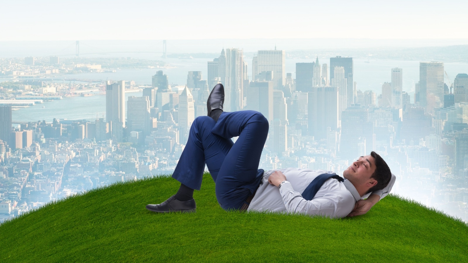 A happy man dressed in business attire resting on a lush green hill in the foreground, while the city waits in the background to represent sabbatical leave.