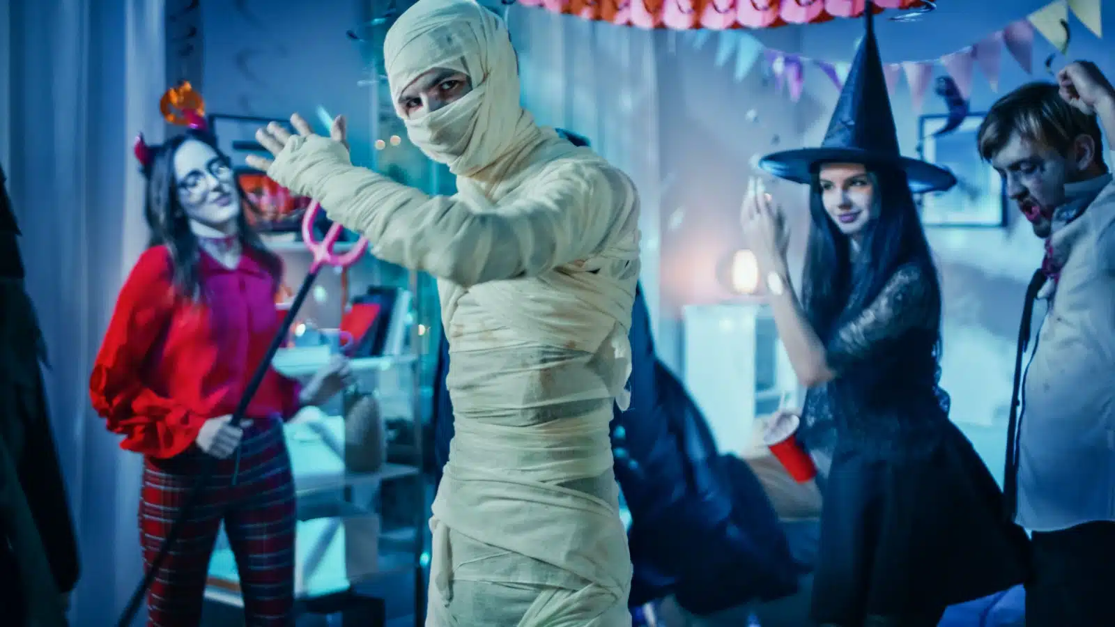 People dressed in traditional monster costumes like a mummy, witch, and zombie dance at a Halloween party. 