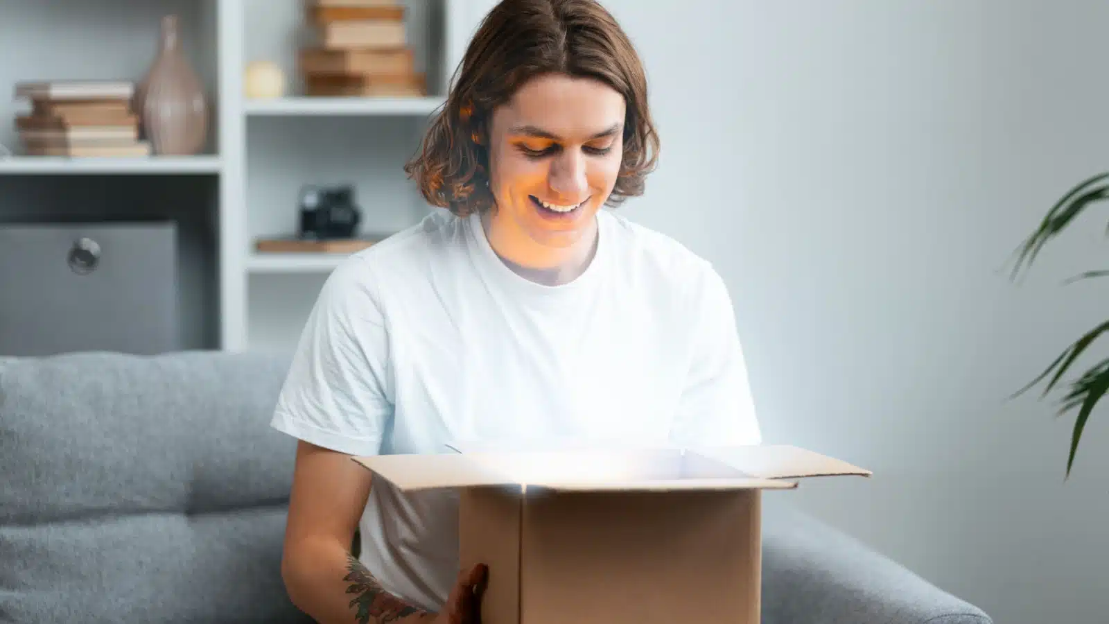 A happy man sits on the couch opening a mystery box that glows.