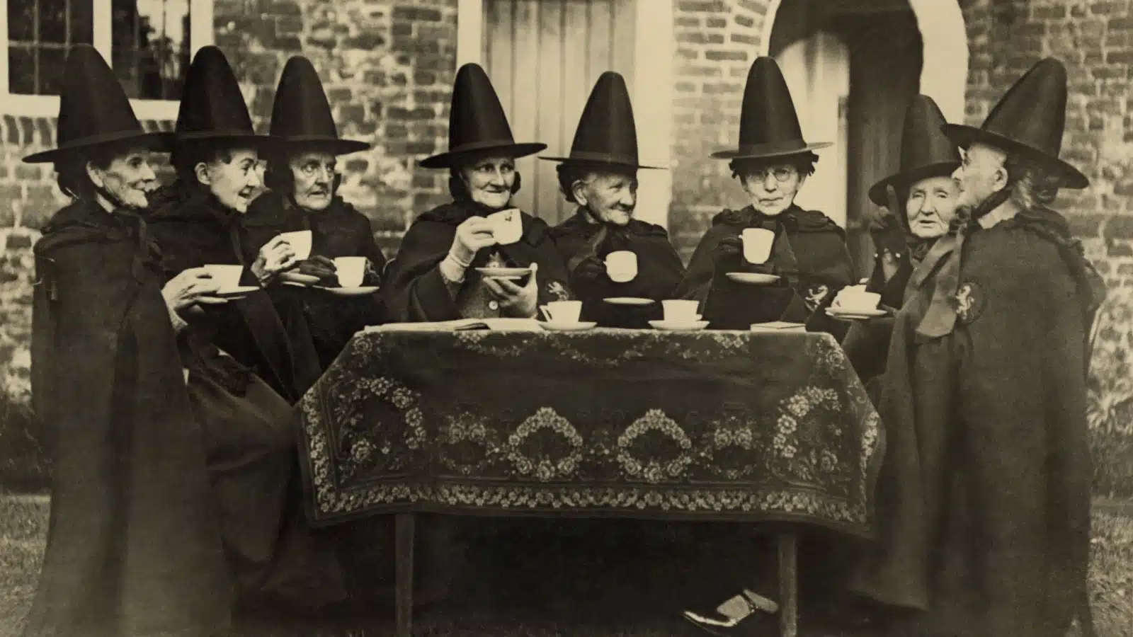 A historical photo of Eight women in high hats having tea. They are not a group of witches but the members of the Holy and Undivided Trinity of Castle Rising, Norfolk, England. Ca. 1920