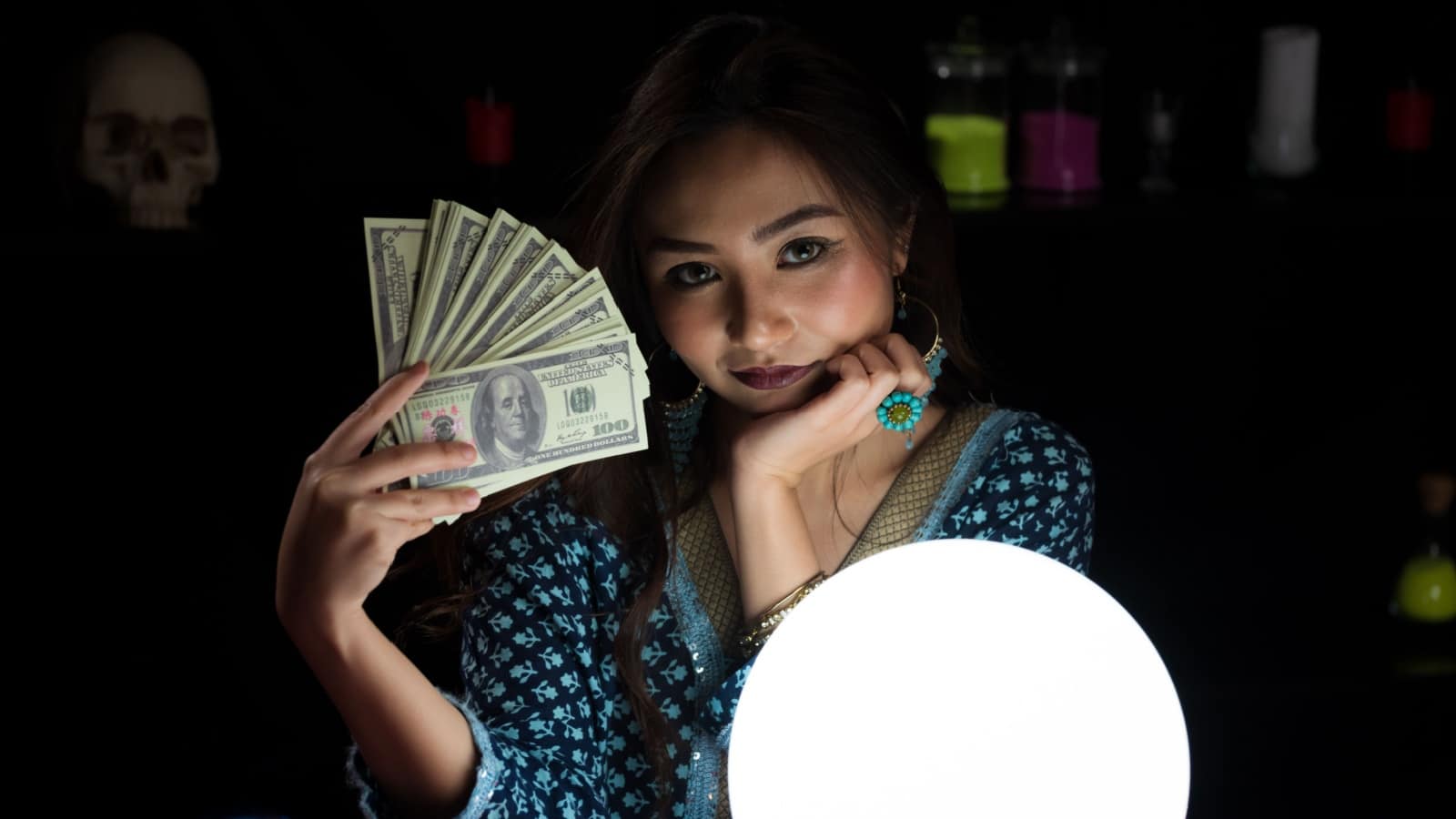 A modern witch sitting in front of a crystal ball holding a fan of money to represent how to cast a prosperity spell.