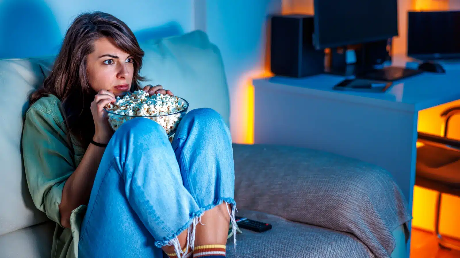 A woman curls into a ball on the couch while eating popcorn and watching a scary movie.