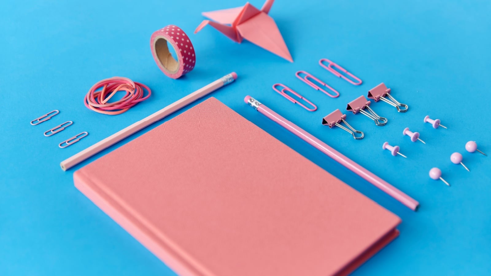 A pink journal surrounded by pink office supplies on a blue background to represent bullet journal supplies.