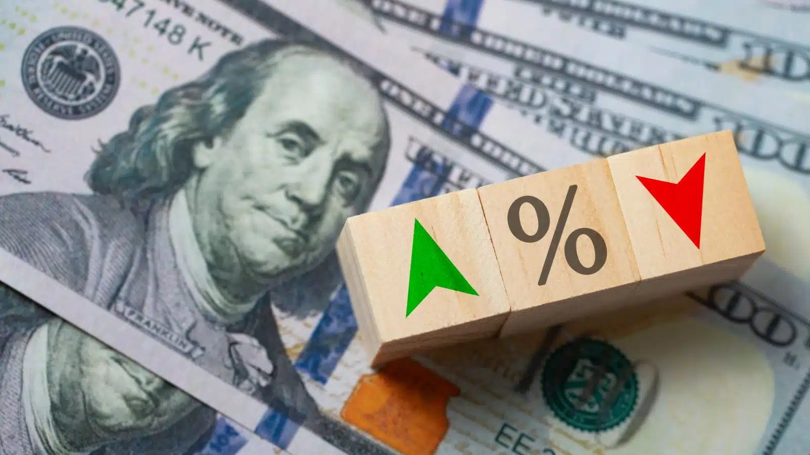 Wooden blocks featuring a green up arrow and a red down arrow next to a percentage sign sitting atop a pile of hundred dollar bills to represent changing interest rates. 