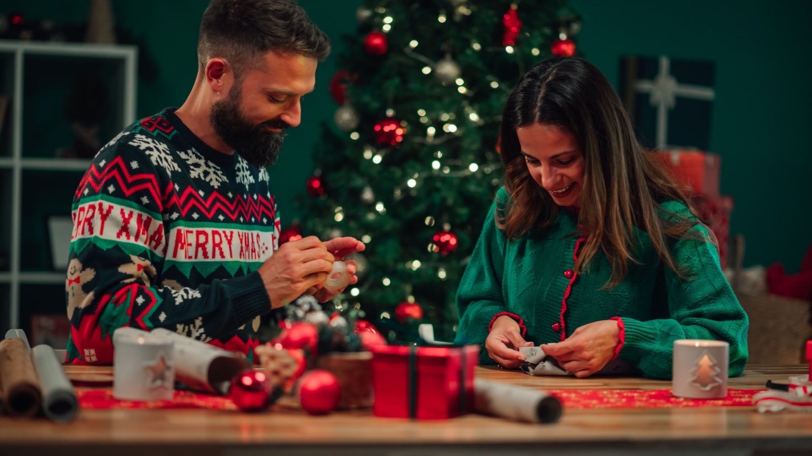 A couple wearing the traditional Christmas colors of red and green sit at a table in front of a Christmas tree wrapping gifts.