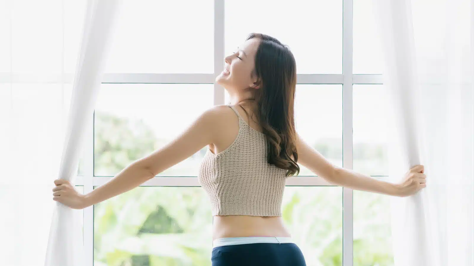A happy energized woman stands at the bright window in early morning.