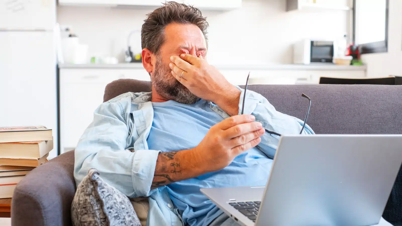 A frustrated man rubs his face while sitting on a couch with his laptop. 