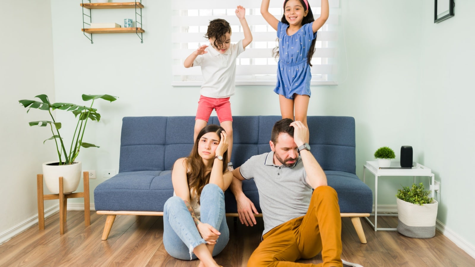 Happy kids jumping on a sofa while tired parents sit on the floor.