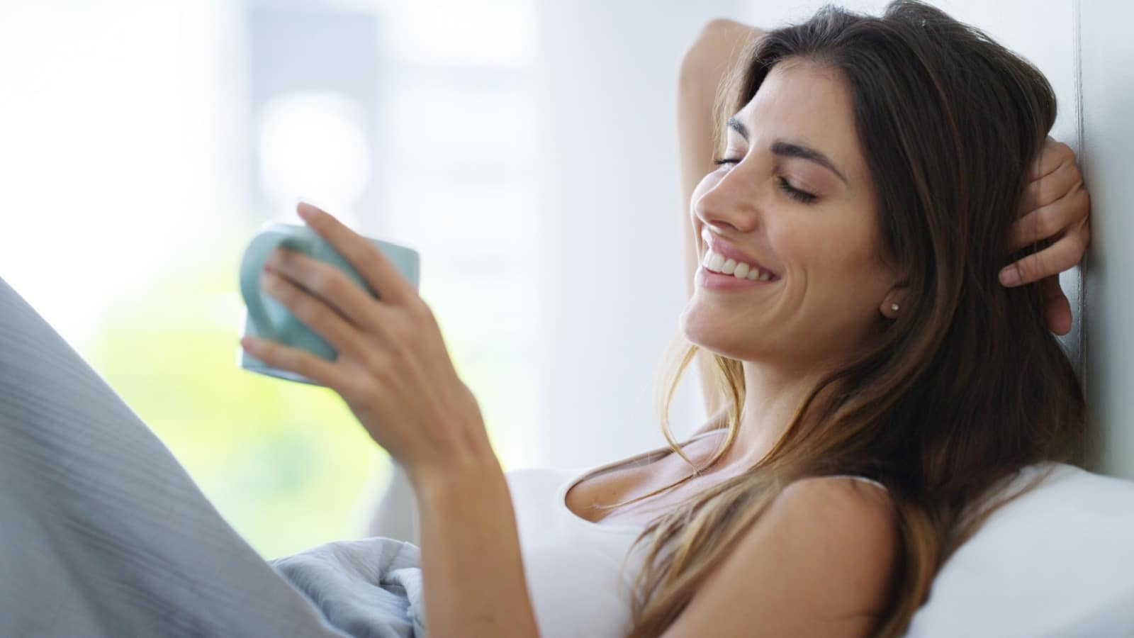 A smiling woman relaxes in bed with a cup of coffee before starting the day to represent the best morning routine ideas.
