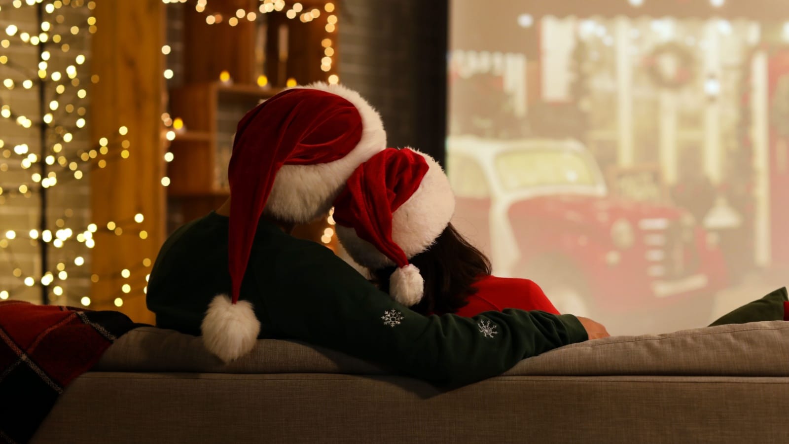 A couple snuggling on the couch wearing Santa hats and watching Christmas movies.