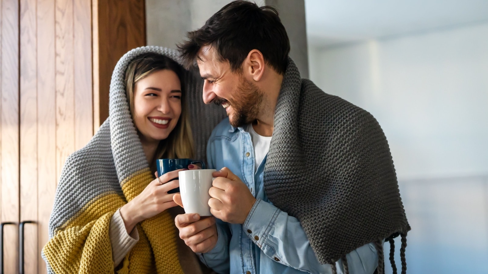 A smiling couple drinks coffee while wearing blankets to represent how to save money on heating.