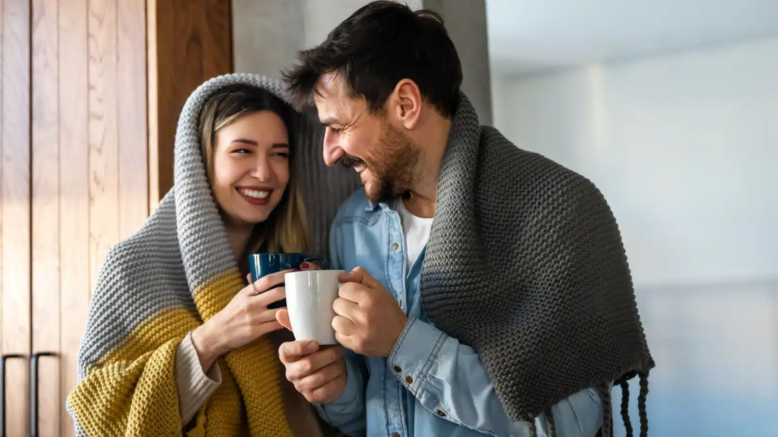 A smiling couple drinks coffee while wearing blankets to represent how to save money on heating.