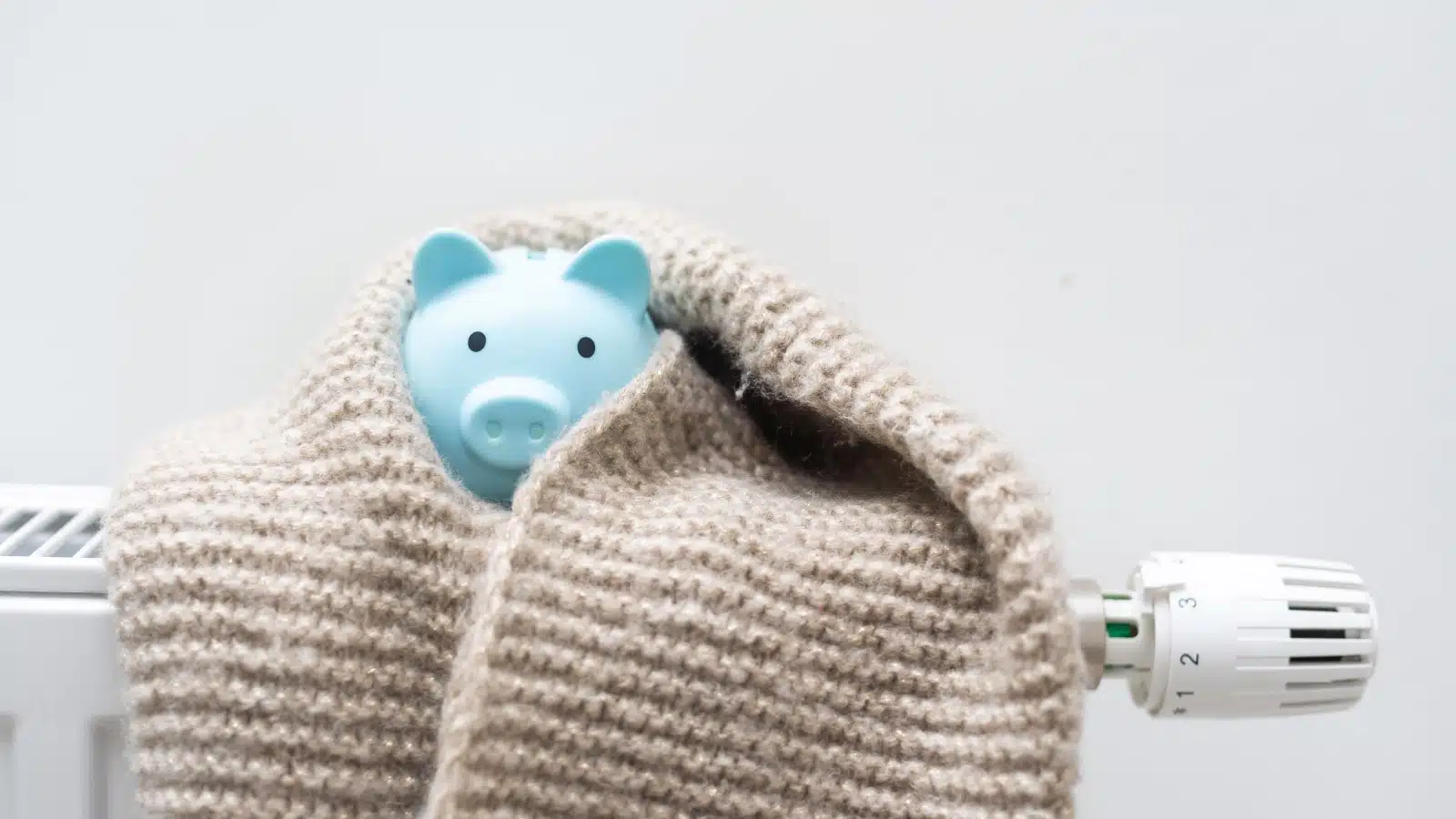 A piggy bank wrapped in a scarf resting on a radiator represents how to save money on heating. 