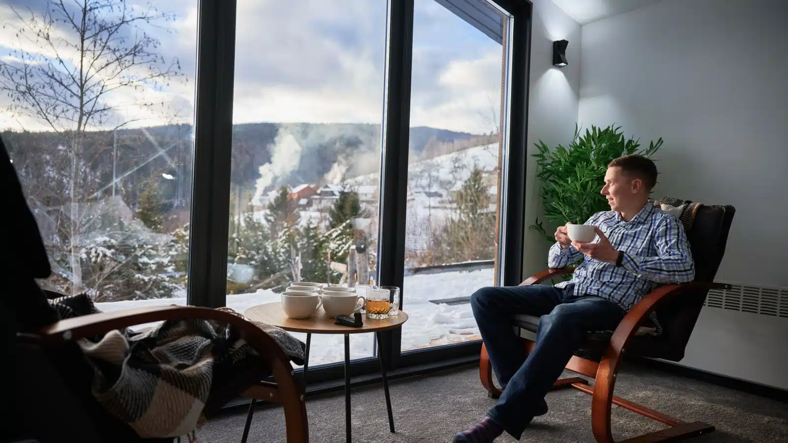 A man sits by massive windows, enjoying a winter's day from inside a warm room.