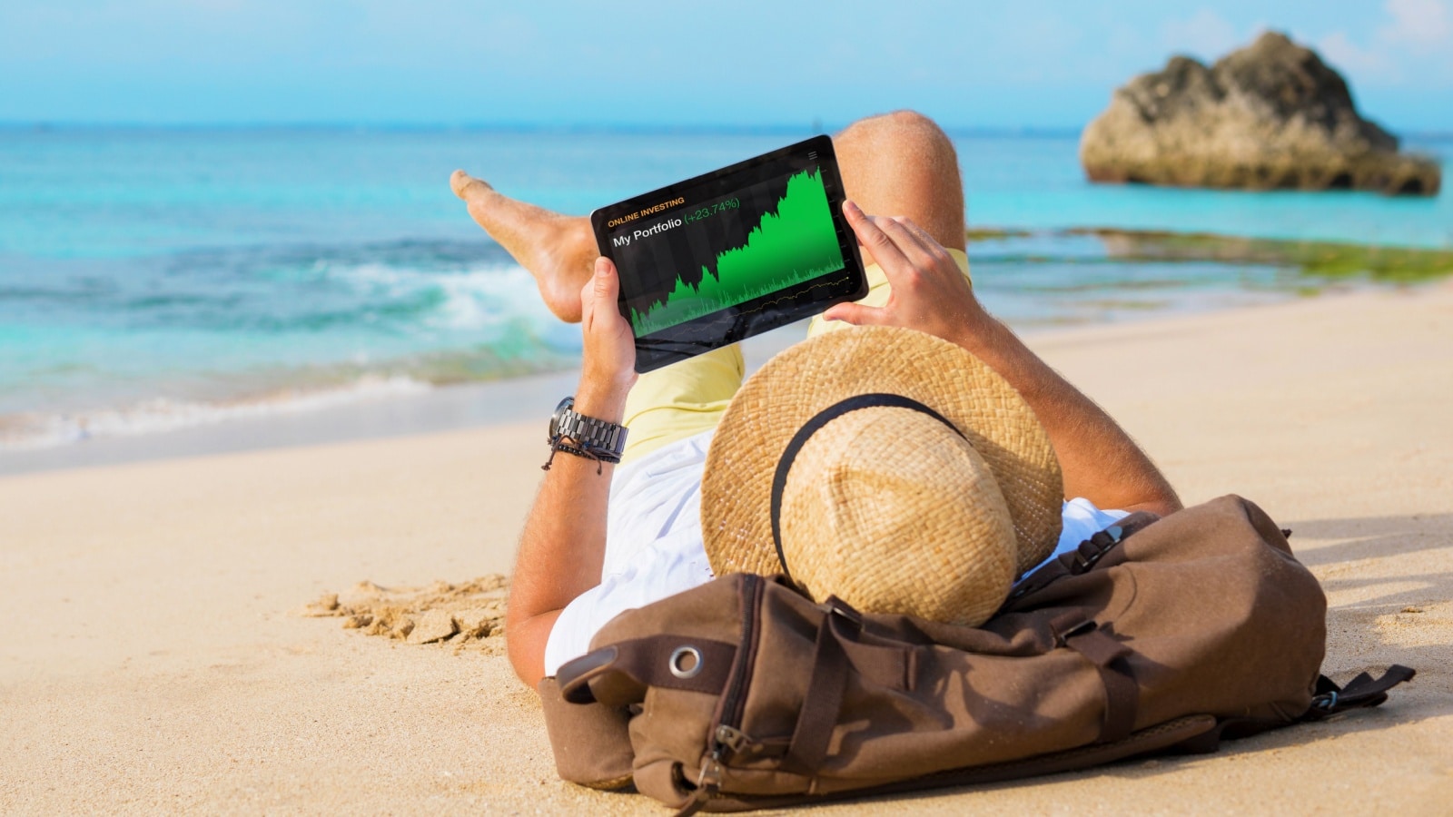 A man checks his investments while lounging on the beach to represent financial freedom.