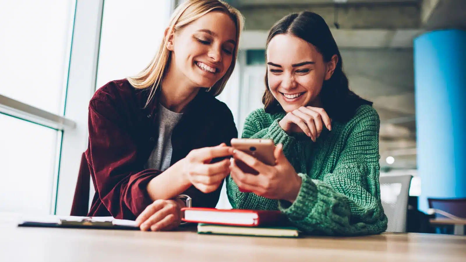 Happy Friends helping each other in a library. Both girls are smiling as they look at something on a phone.