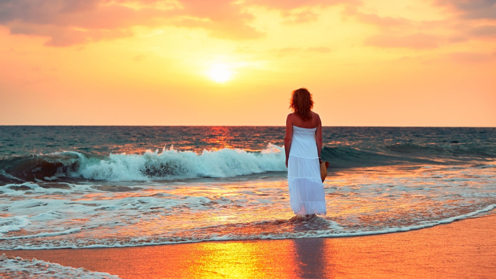 A woman stands by herself on the beach at sunset, to represent the top life experiences everyone needs to have at least once.