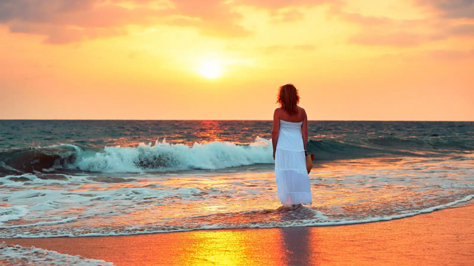 A woman stands by herself on the beach at sunset, to represent the top life experiences everyone needs to have at least once.
