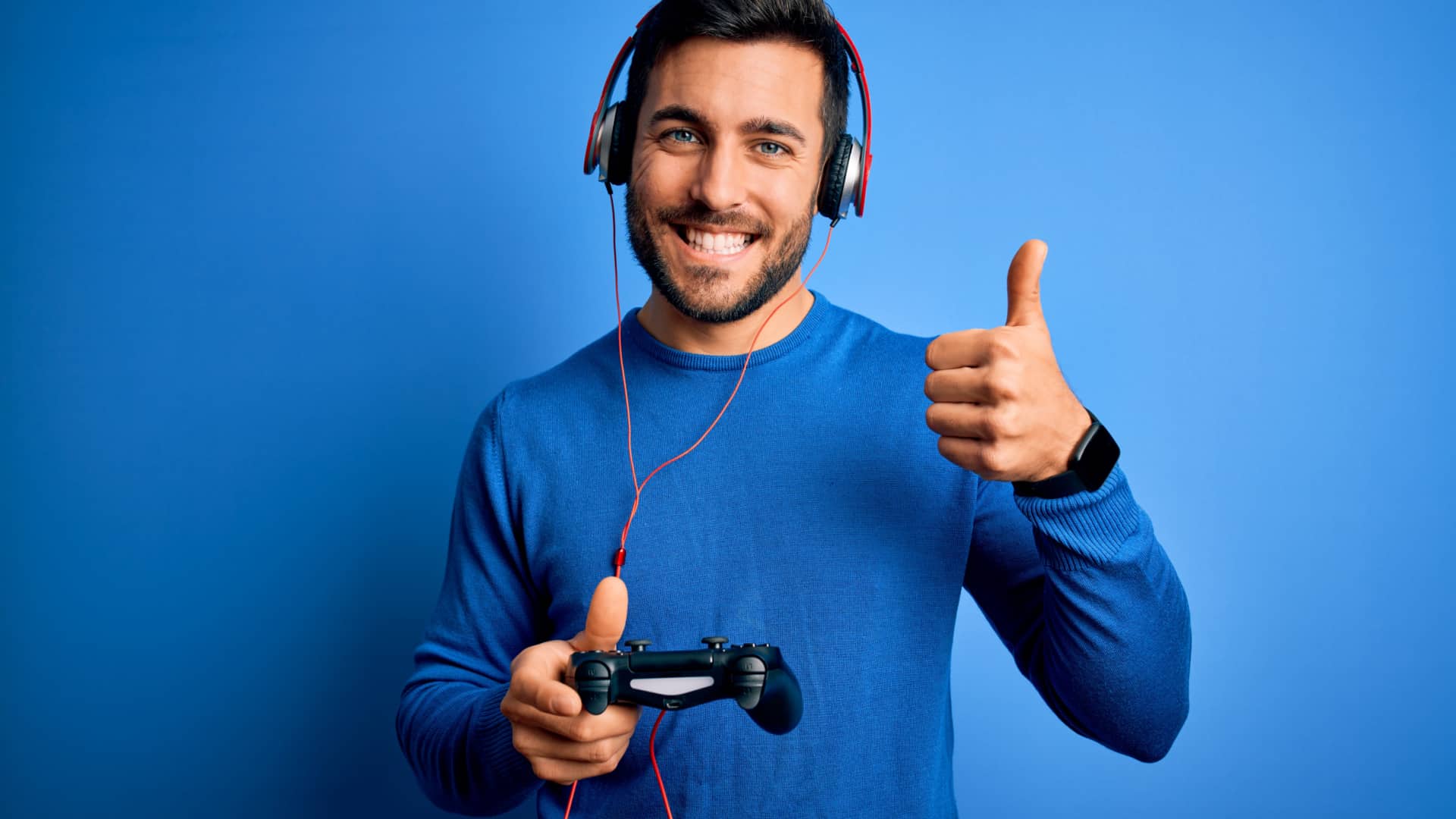 man wearing blue on blue background holding video game controller wearing headphones and smiling while giving a thumbs up