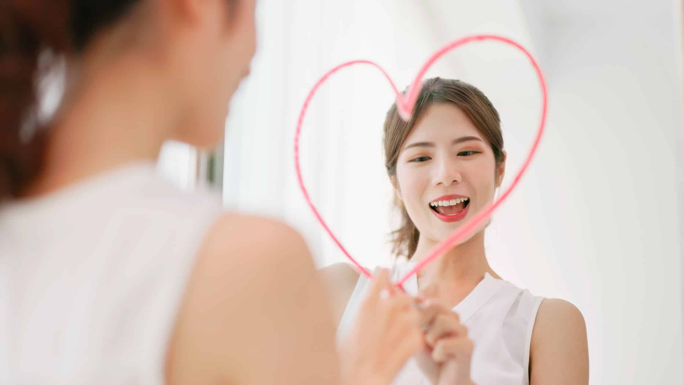 Woman looking at herself in a mirror drawing a heart over her reflection to represent shadow work