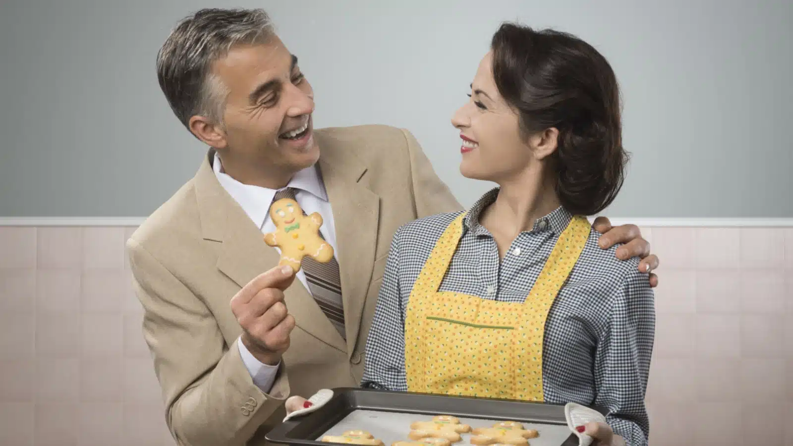 Vintage looking photo of a 1950s husband and wife. The husband is dressed for work while the wife offers freshly baked cookies.