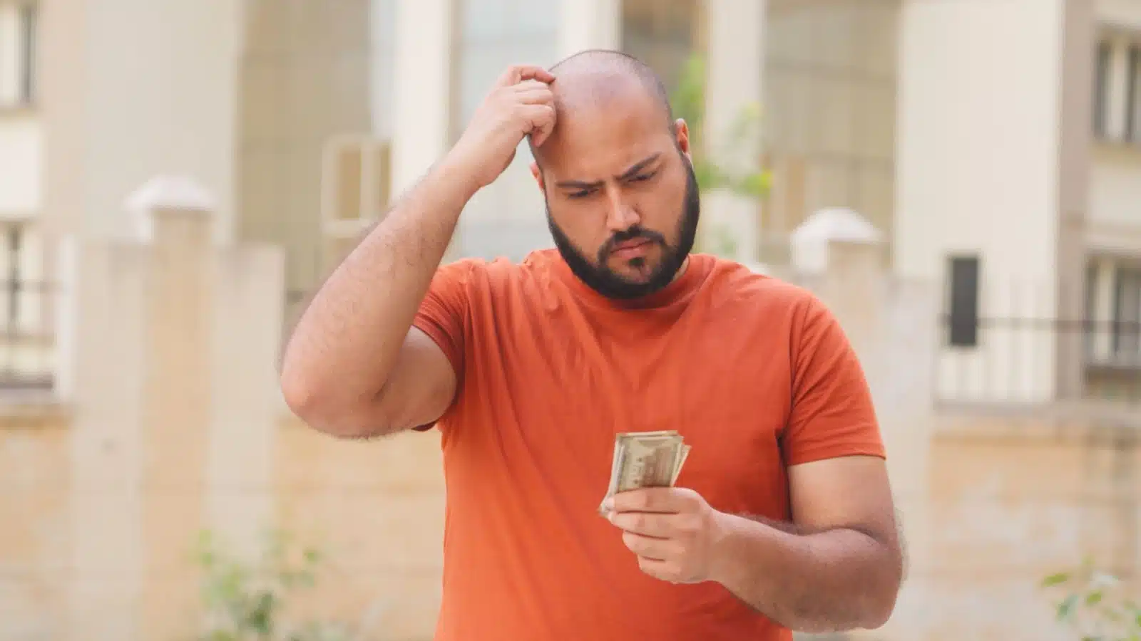A confused looking man scratches his head while looking at his money.