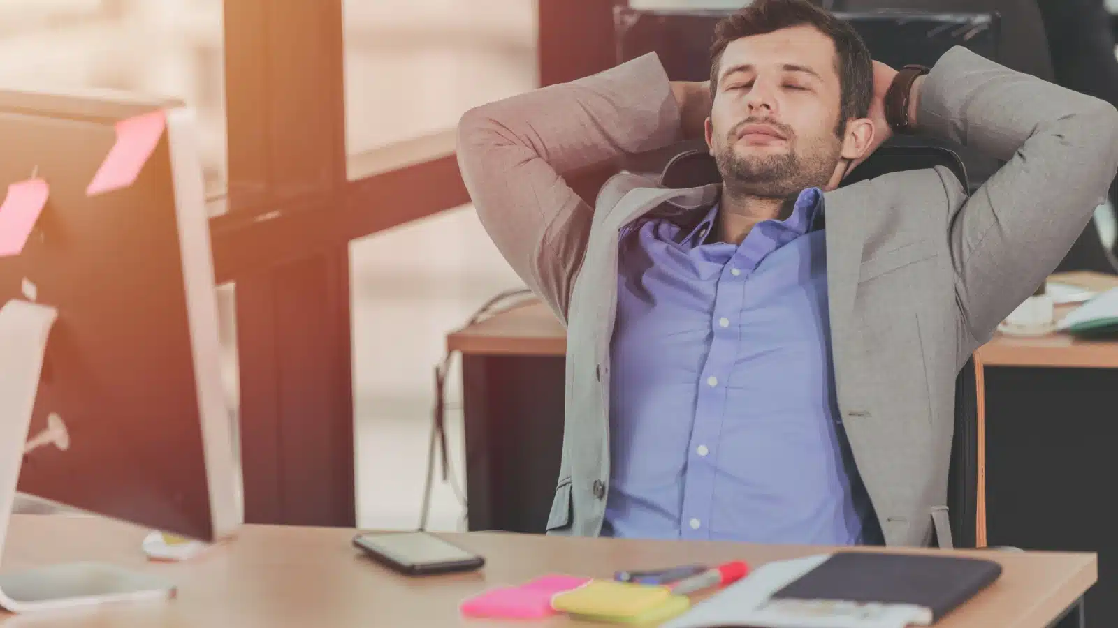 A man leans back and closes his eyes while working at his desk.