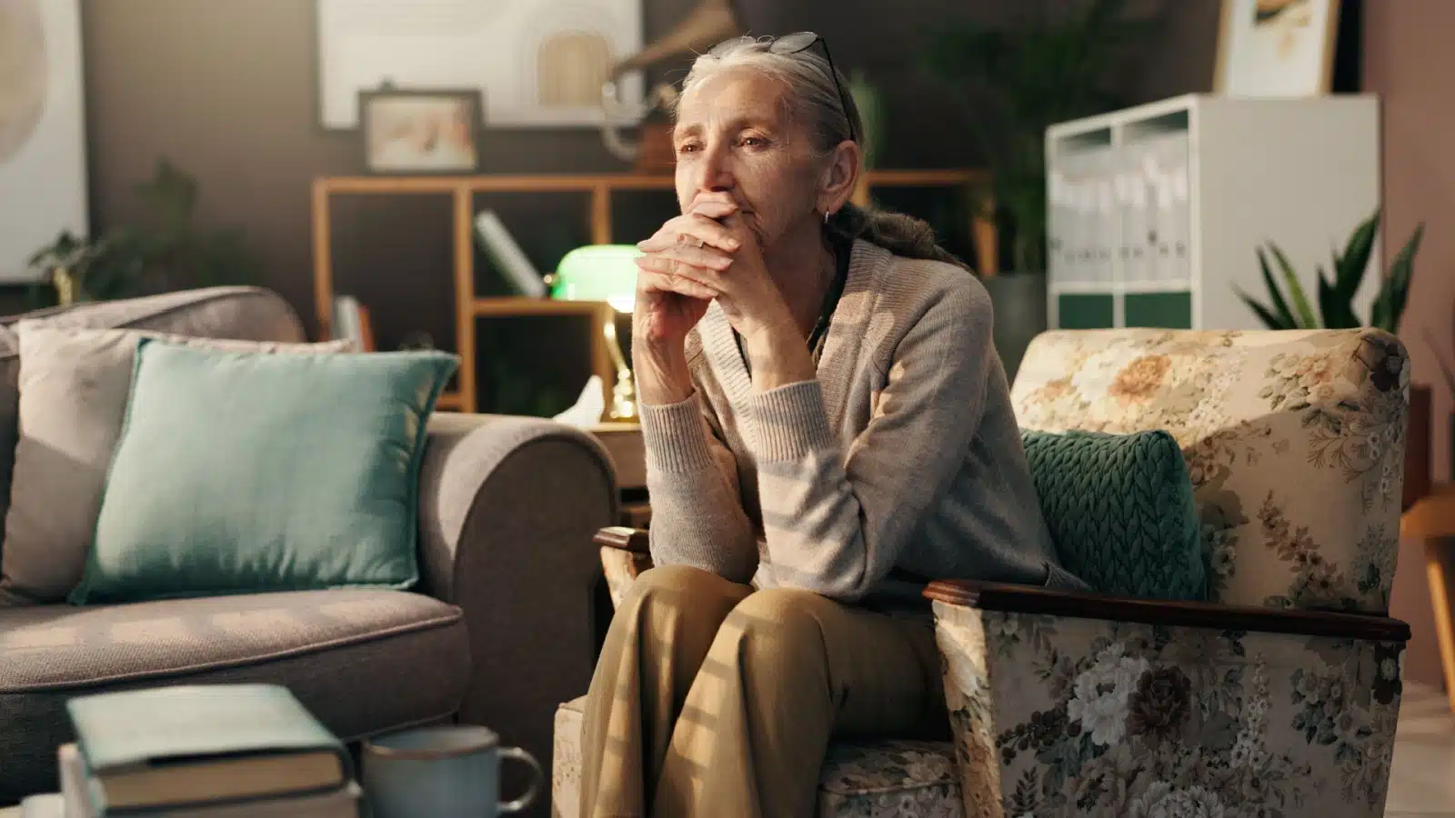 An older woman sits alone in her living room, looking thoughtful and pensive. 