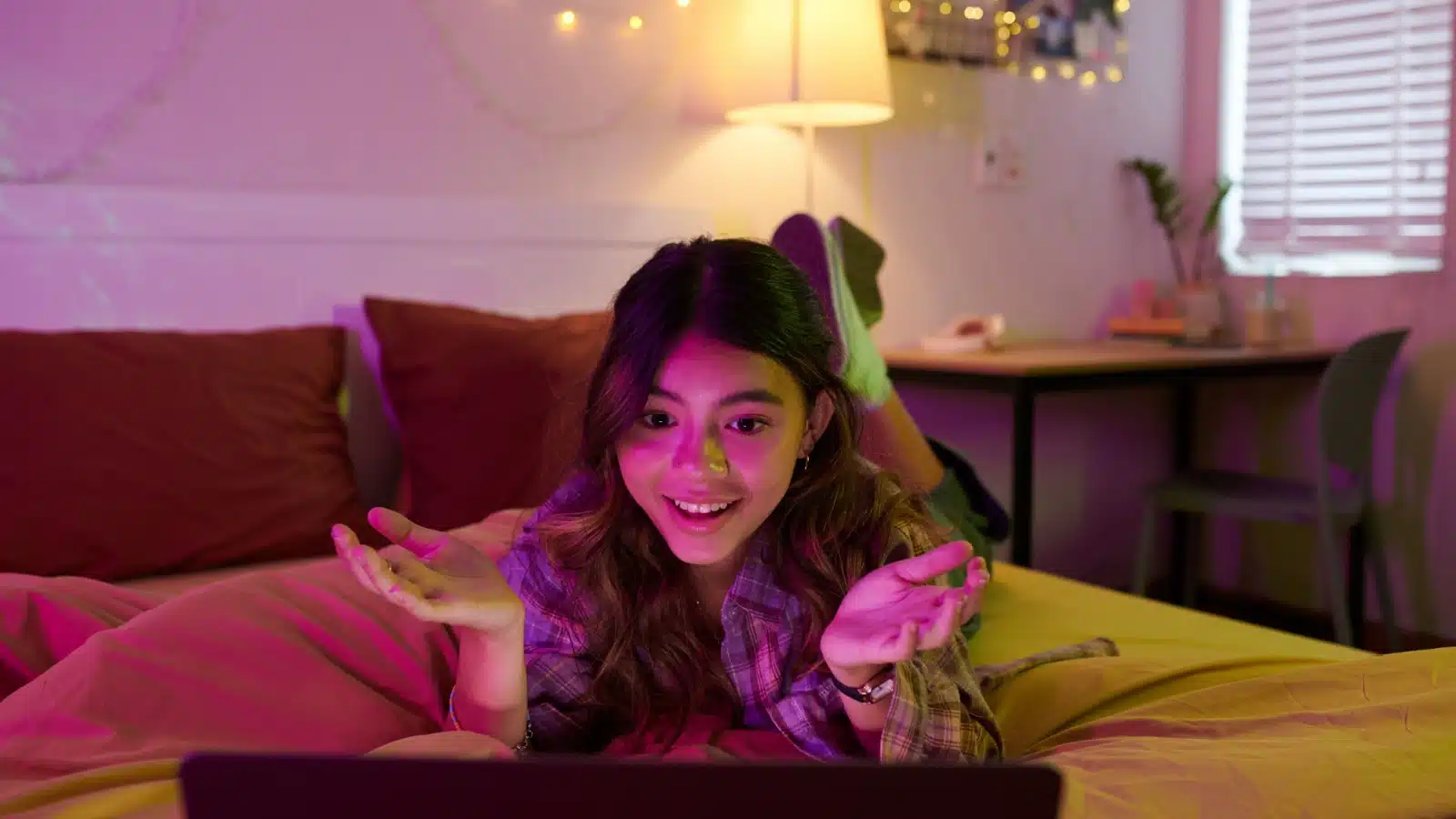 A teenager lies on her bed, talking with her friend on a video call with her laptop. 
