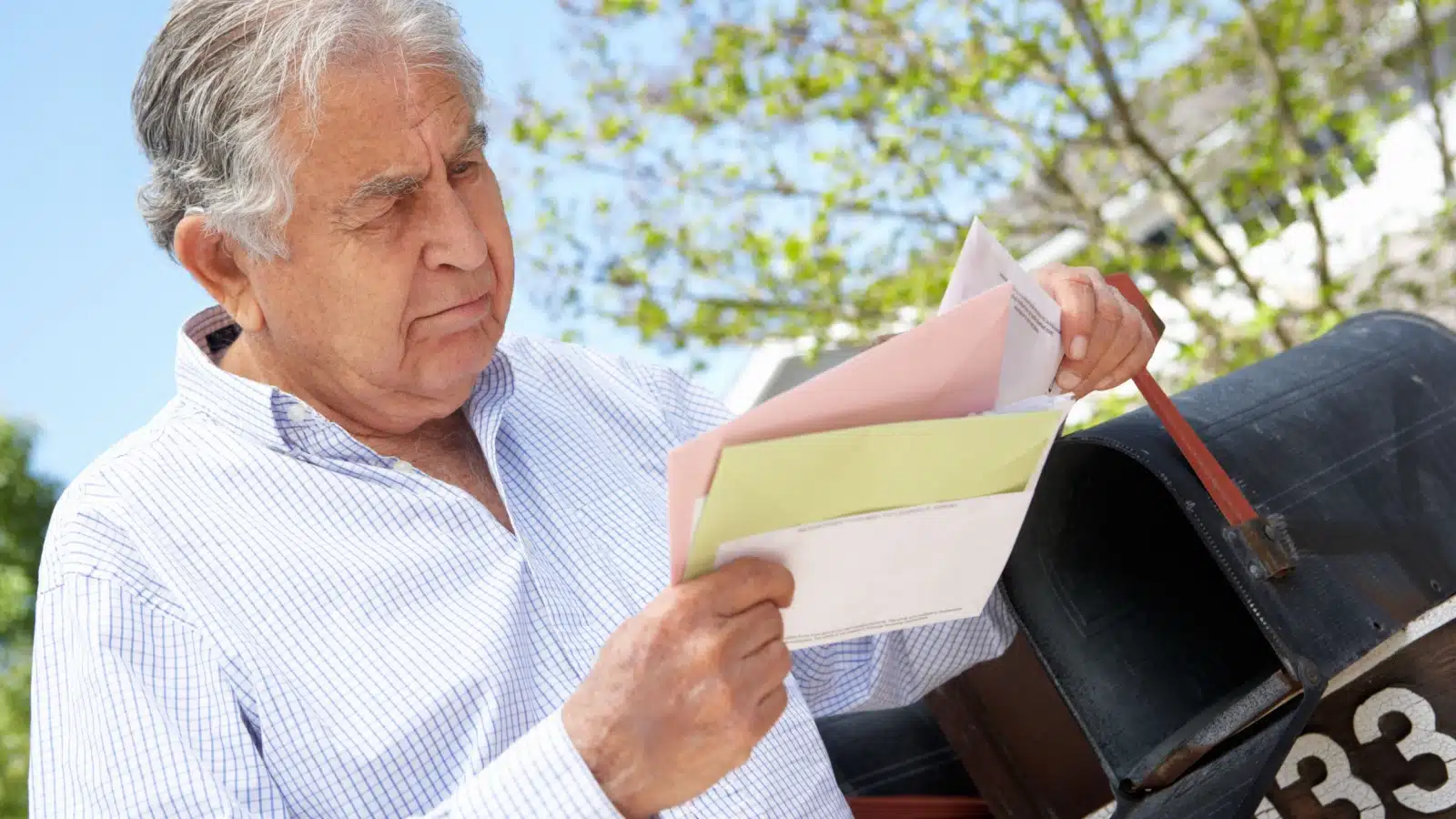 A grumpy looking older man stands by his mailbox checking his mail.