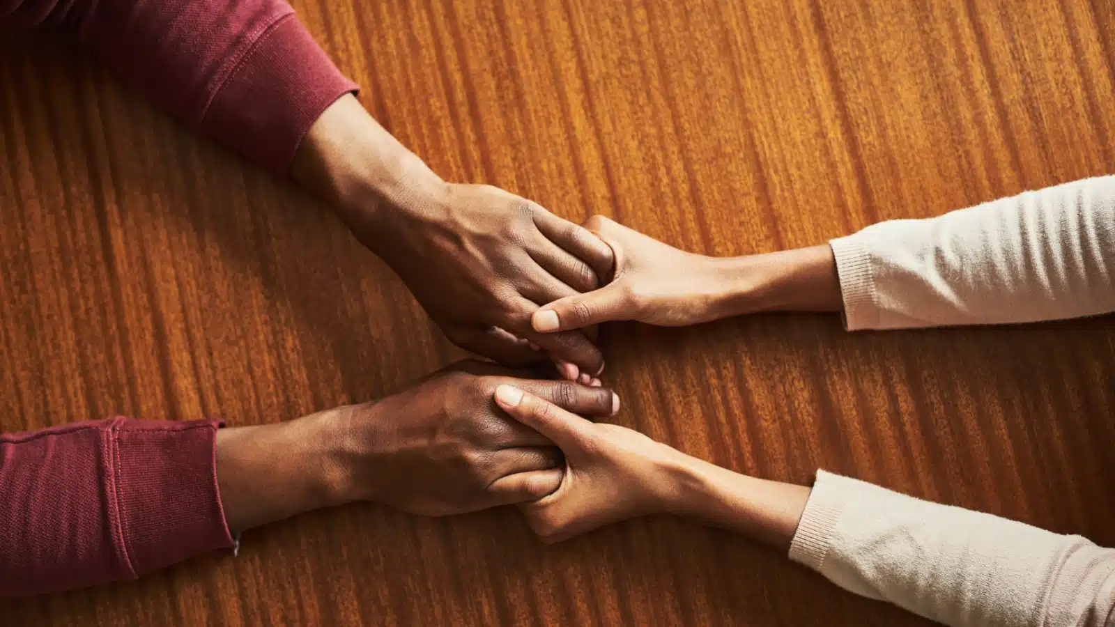 People holding hands from across a wooden table. The image only shows the arms and hands.