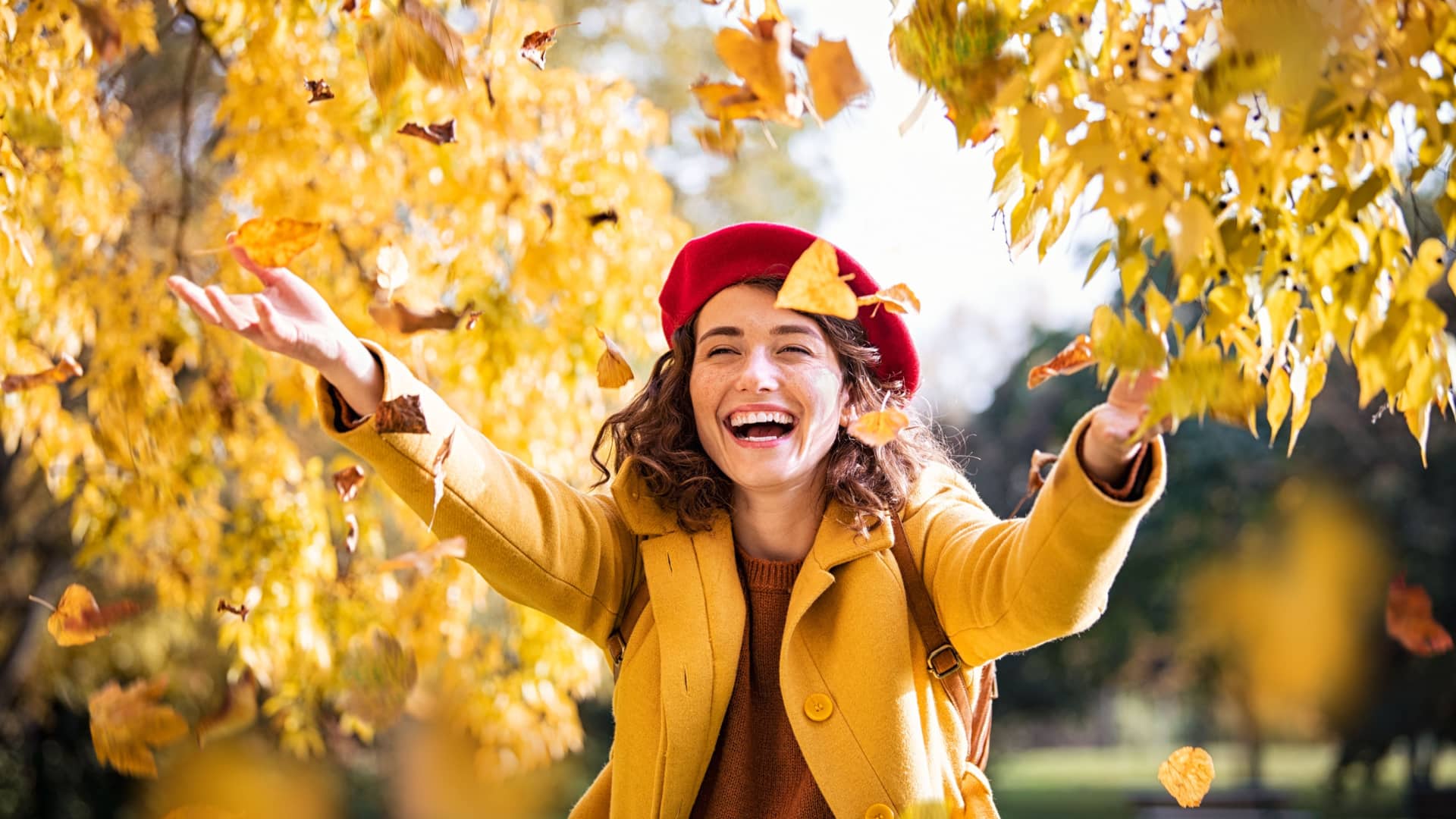 woman smiling while walking through autumn trees with yellow leaves falling around her to represent fall activities