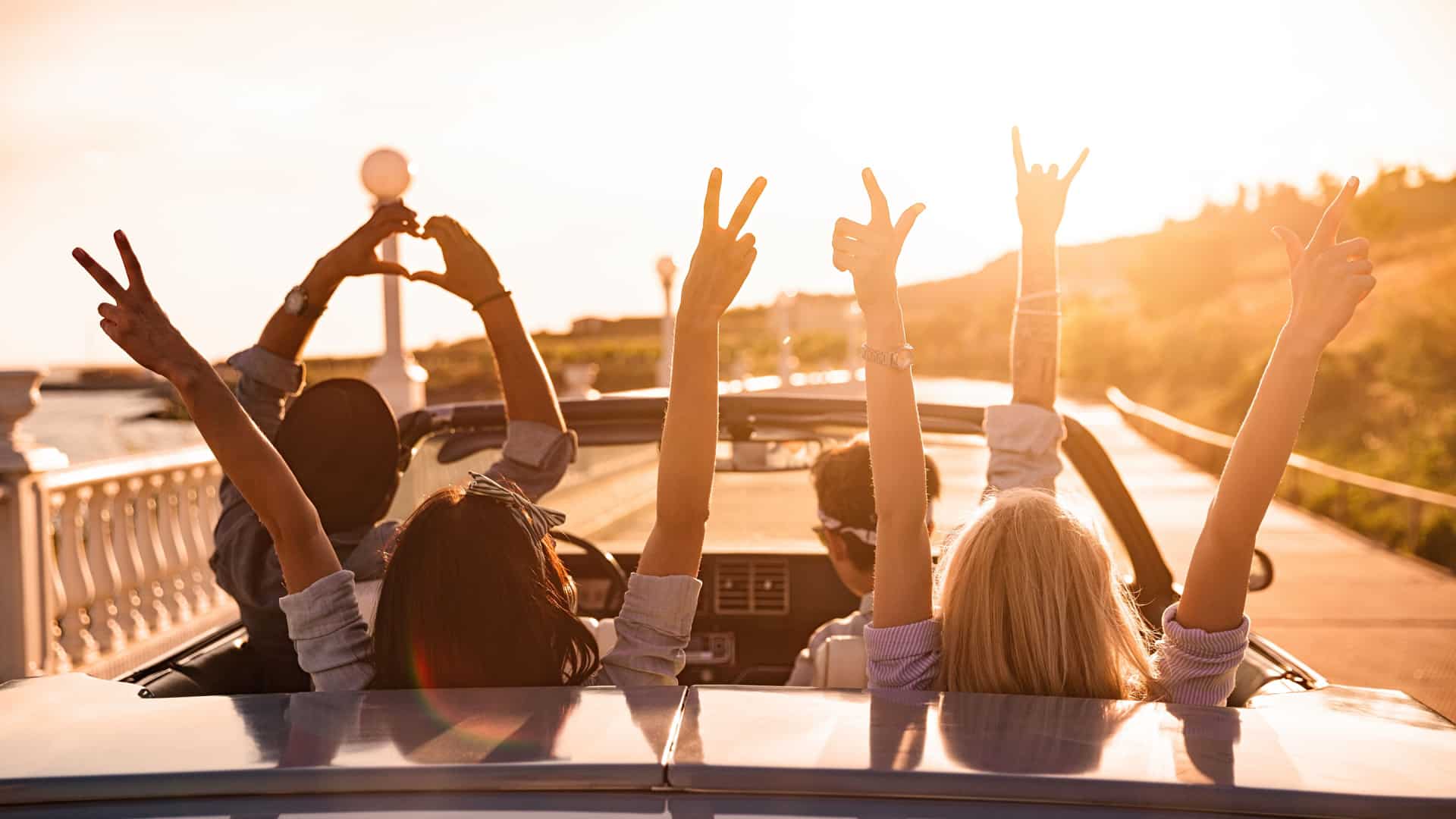 Group of young adults riding in a convertable with their hands up