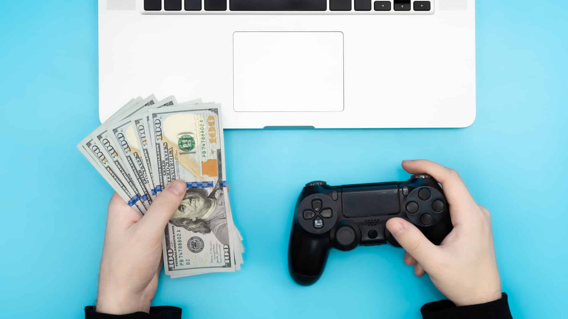 hands holding cash and a video game controler in front of a laptop to represent making money gaming
