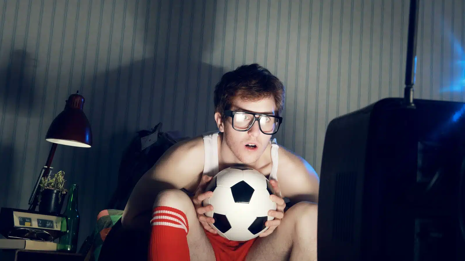 A man is hunched over in a dark room, cuddling his soccer ball, obsessively starting at the screen. He takes his love of sports too far.
